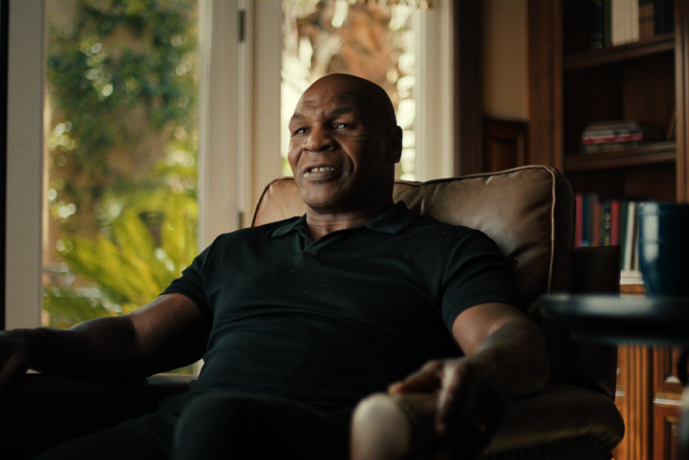 "A man sitting in a cozy armchair in a warmly lit home, surrounded by bookshelves and large windows with sunlight and plants visible outside, a coffee table with a mug in the foreground."